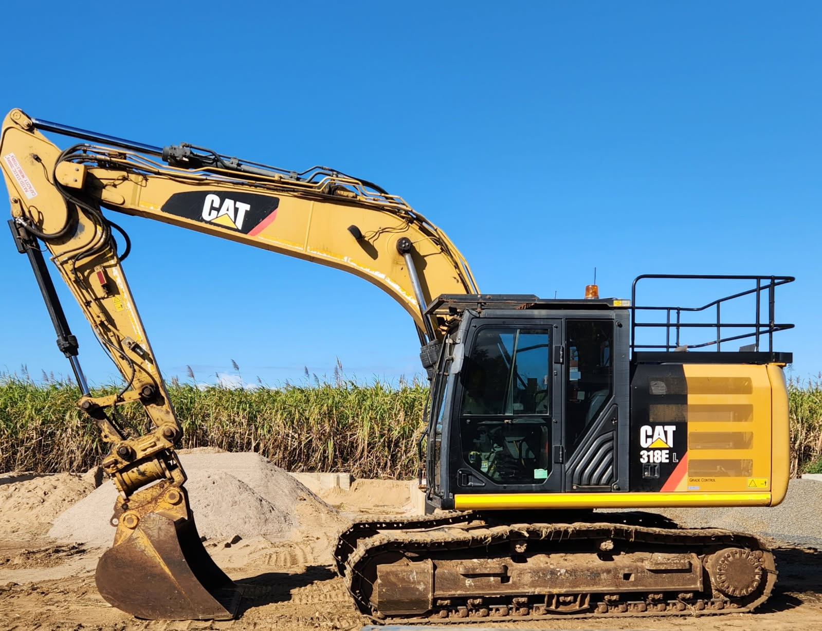 A yellow CAT 318E excavator is parked on a construction site with dirt piles and tall grass in the background under a clear blue sky.