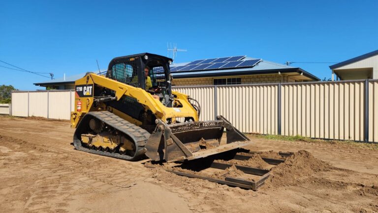 A yellow Caterpillar compact track loader moves dirt on a construction site near a beige fenced house with solar panels on the roof.