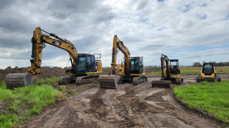 Four yellow excavators of varying sizes are parked on a dirt construction site with grassy areas and a cloudy sky in the background.