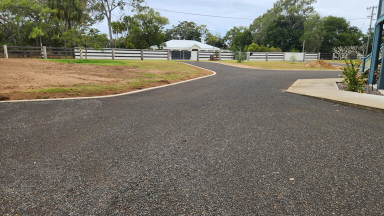 A newly paved asphalt driveway, shaped by expert residential earthworks, curves toward a white house, with fenced grassy areas and trees in the background under a cloudy sky.