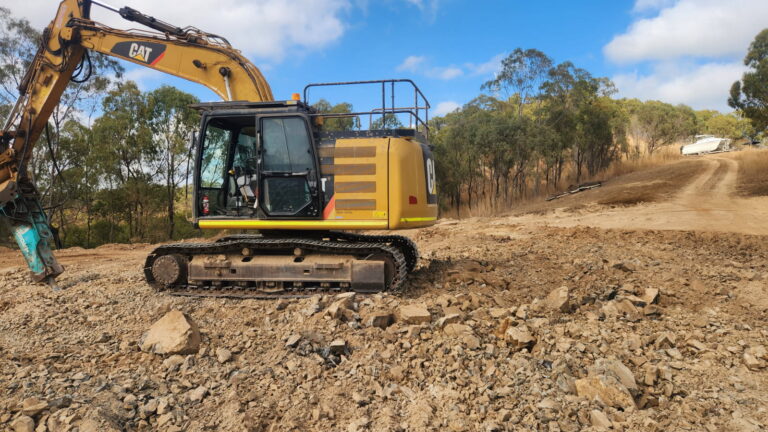 A yellow excavator is parked on a rocky dirt construction site, with trees and a dirt road visible in the background under a partly cloudy sky.